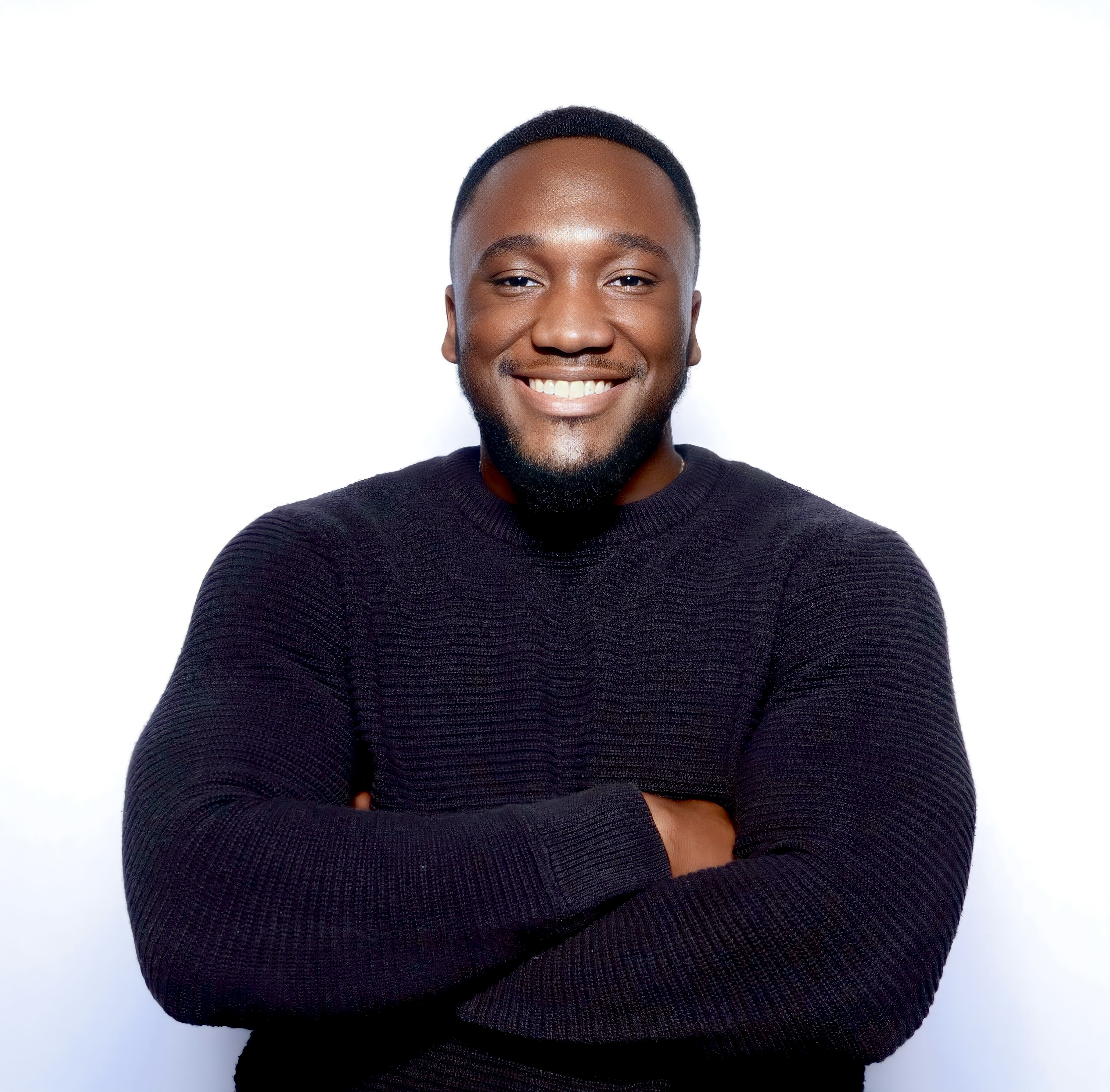 Black man with short hair and a fade wearing a black sweater, sitting in a brightly-lit; he is looking at the camera with a huge smile on his face.