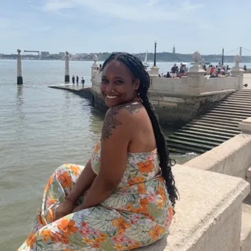 Black woman sitting on the docks near a large body of water and looking over her shoulder, smiling; she has a tattoo covering her shoulder, long braided hair, a nose ring, and a floral-patterned dress.